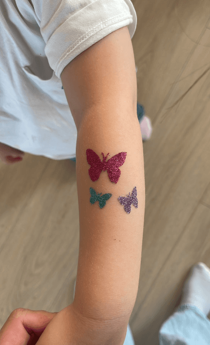 Drie glitter-tattoos in de vorm van vlinders op een onderarm: roze, groen en paars
